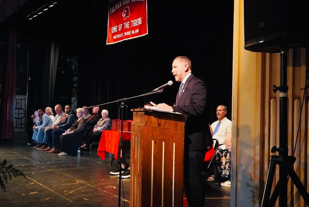 Juan Pablo Barrera speaking at a podium on a school auditorium stage.